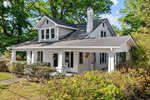 A quaint one-and-a-half-story house with a covered porch, surrounded by greenery on a sunny day.