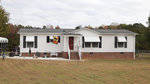 A single-story white modular home with a red door and a few steps leading up to the entrance. the house has a dark gray roof, and there is a flag displayed on a pole in the front yard. the property is bordered by a thin wire fence and includes a carport on the left side. the surrounding grass lawn appears to be well-maintained.