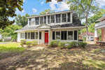 A two-story suburban house with white siding and black shutters, featuring a red front door and surrounded by trees and a lawn.