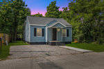 A small blue single-story house with white trim stands at twilight, featuring a gravel driveway and a concrete sidewalk leading to the front steps, surrounded by trees and vegetation.
