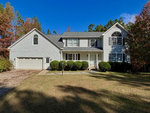 A two-story suburban house with a white exterior, two-car garage, and a front yard with a grass lawn and a few trees under a clear sky.