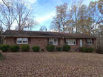 A single-story brick house with a dark roof surrounded by leaf-strewn lawn and bare trees, indicating autumn or winter season.