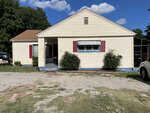 A single-story beige house with red shutters, a front porch, a driveway on the right side, and two small bushes flanking the front steps, under a clear blue sky.