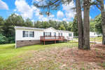 A single-story manufactured home with white siding and a red wooden deck, set against a backdrop of trees and a clear blue sky.