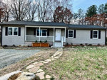 A single-story suburban house with gray siding, white trim, a small front porch, and a stone walkway leading to the front door, surrounded by a grassy lawn with sparse landscaping and a backdrop of bare trees.