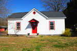 A small white house with red trim and a red door, situated under a clear blue sky.