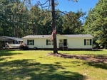 A single-story light green house with white trim under a clear sky, featuring an american flag on a flagpole, a front yard with a tree, and a carport on the left side.