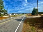 A rural road stretches ahead flanked by grassy verges, power lines, and sporadic buildings under a cloudy blue sky.