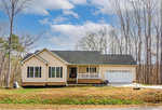 A single-story beige house with white trim and a front porch, featuring a two-car garage on a sunny day with a partially cloudy sky and leafless trees in the background.