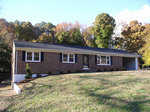 A single-story red brick house with a black shingle roof, featuring white-framed windows, and a front lawn with young trees and shrubs under a clear sky with a few clouds.