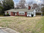 A single-story brick house with white siding and window shutters, featuring a chimney and a carport, situated on a lawn with a small patch of visible bare ground and a partly cloudy sky overhead.