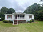 A single-story white house with red shutters, featuring a front porch with stairs, surrounded by a lawn and trees under an overcast sky.