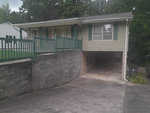A single-story house with green siding, featuring a raised front porch with green railings, a stone retaining wall, and an under-house garage.
