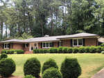 A single-story brick house with white-trimmed windows, surrounded by a manicured lawn and neatly trimmed bushes, with a backdrop of tall green trees.
