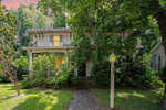 A two-story house with white siding partially obscured by leafy green trees, featuring a front porch and a brick pathway leading to the entrance, under a serene sky at dusk.