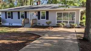 A single-story house with white siding and dark shutters, featuring a small front porch with wooden stairs, surrounded by a freshly mulched garden bed, and a concrete walkway leading to the front door.