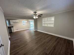 An empty room with wooden floor, a ceiling fan, and an open-plan kitchen in the background.