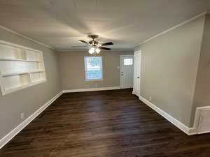 An empty room featuring dark wood flooring, white walls with one accent wall containing built-in shelving, a ceiling fan, and a closed door with a window to the outside.