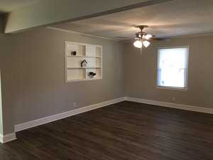An empty room with beige walls, dark laminate flooring, a ceiling fan with lights, and a built-in white shelving unit with a few decorative items on the shelves. a window provides natural light to the space.
