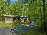 A single-story residential home with a dark roof and light-colored siding, featuring an attached garage, is surrounded by tall, lush green trees. a paved driveway leads to the garage and a set of stairs leads to the main entrance.