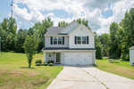 A two-story detached house with a white exterior and gray roof featuring an attached two-car garage and a concrete driveway, set against a backdrop of a lawn with scattered trees and a partly cloudy sky.