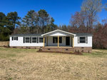 A single-story house with white siding and brown brick foundation, featuring a central covered entrance with two yellow pillars, set on a grassy lawn with trees in the background under a clear blue sky.
