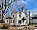 A two-story white house with black shutters, a front porch, and bare trees in the yard under a partly cloudy sky.