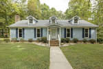 A single-story blue suburban house with a gabled roof, white trim, a central door, and symmetrically placed windows, surrounded by greenery and a front lawn.