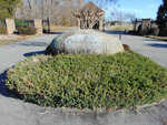 A landscaped entrance area featuring a large rock with the inscription "peninsula" resting on a bed of low-lying shrubbery, with metal gates visible in the background under a clear sky.