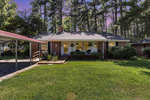 A single-story suburban house with a brick facade, white trim, and a covered front porch, surrounded by tall pine trees and a well-maintained lawn.