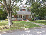 A quaint suburban brick house with a white porch under a large tree, featuring a well-kept front lawn and a clear sky above.