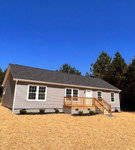 A single-story house with beige siding and a small front porch under a clear blue sky, surrounded by a freshly mulched yard with a backdrop of tall trees.