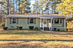 A single-story house with light green siding, a gray shingled roof, and white trim, featuring a small porch with stairs and surrounded by a few trimmed bushes and pine trees.