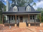 Newly constructed single-story house with a symmetrical front facade, featuring a central front door flanked by two dormer windows and a covered porch with railings, set against a backdrop of trees.