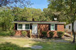 A single-story brick house with white trim windows, a small front porch, and a satellite dish, situated in a leafy area.
