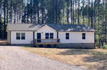 A single-story residential home with white siding and a small wooden front deck, surrounded by pine trees under a clear sky.