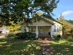 A single-story yellow house with a porch, partially obscured by trees, on a sunny day with a clear blue sky.