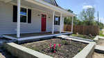 A single-story house with gray siding and a red front door, featuring a wooden porch and garden beds with blooming pink flowers in the foreground.