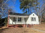 A small white single-story house with a brick chimney and a front porch, surrounded by bare trees under a clear blue sky.