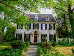 A stately two-story colonial house with a brick facade, white trim, and a central front door, surrounded by mature trees and greenery.