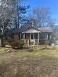 A single-story brick house with a white front door, flanked by windows, nestled among trees with a front yard lacking grass.