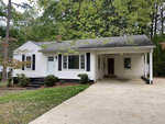 A single-story white house with black shutters and a covered porch surrounded by trees and a driveway on the left side.