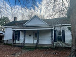 A weathered, white single-story house with dark shutters, a central chimney, and visible signs of neglect, including a sagging gutter and overgrown yard.