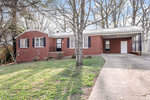A single-story red brick house with white trim, featuring a covered carport, and surrounded by a lawn with bare trees and a metal fence to the side.