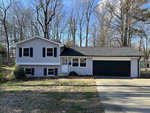 A split-level house with white siding and a black roof features an attached two-car garage, situated in a neighborhood with trees and a clear blue sky overhead.