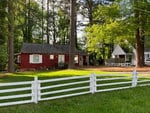 A quaint red house with white trim sits behind a white picket fence, surrounded by lush greenery in a tranquil neighborhood.