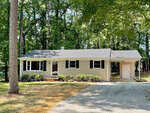 A single-story beige house with a white door, surrounded by trees and featuring a carport on the left side.