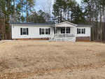 Single-story white house with front porch and white railing, set on a wide lawn with patchy grass, surrounded by trees with no leaves, suggesting late fall or winter season.