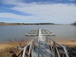 A narrow metal walkway leading to a series of empty boat docks on a calm lake, with clear skies and a treeline in the background.