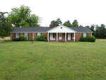 A single-story brick house with white trim and a central entryway, set back from a large front lawn with scattered trees and shrubs.